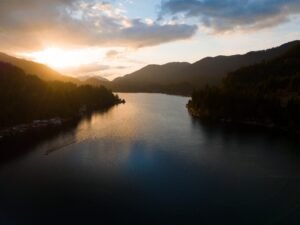 Aerial over a calm lake between forested ridges with the sun setting behind hills
