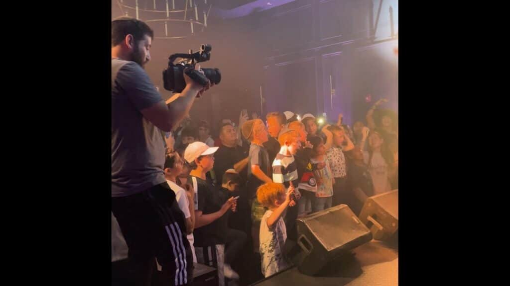 Operator on a stage filming a crowd with raised hands under hazy concert lighting