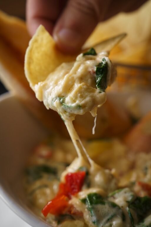 Hand dipping a tortilla chip into cheesy spinach dip with red peppers