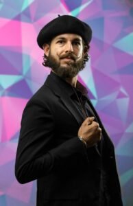 Bearded man in a black beret and black blazer against a pink and teal geometric mural