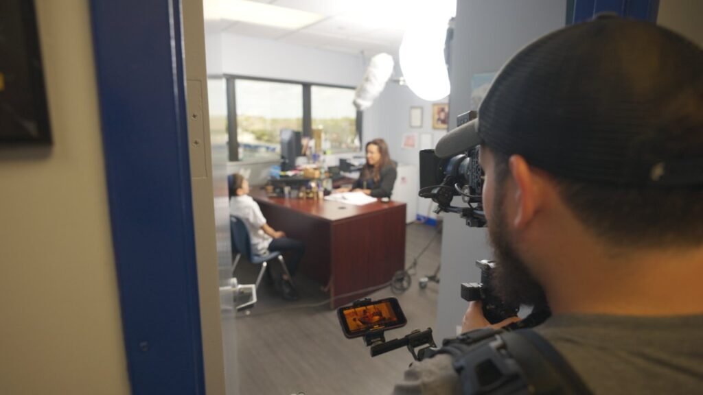Camera operator in a cap filming three people at an office desk through a doorway