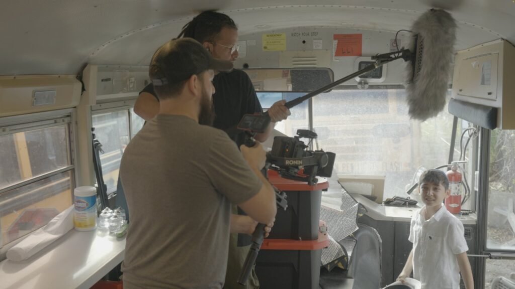 Film crew with camera and boom microphone shooting inside a school bus, child standing at right