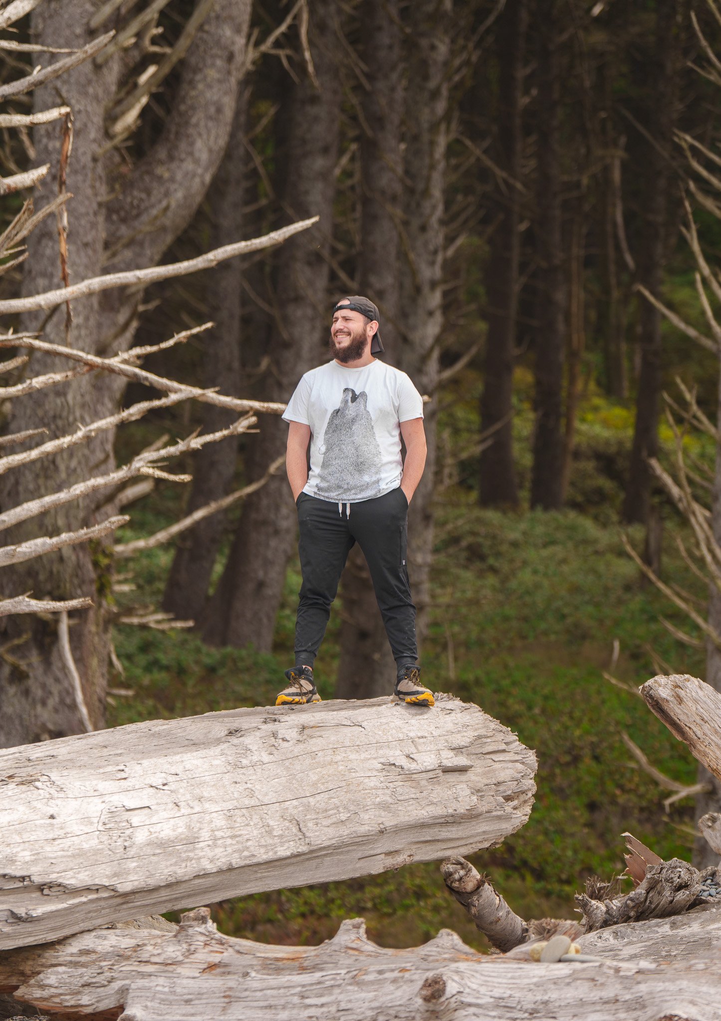 Bearded man in a white shirt standing atop a large fallen log in forest