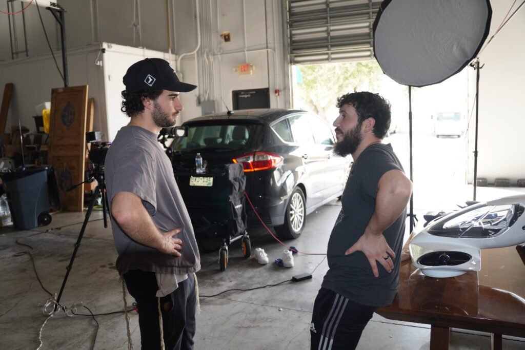 Two men talking in an open garage beside a camera rig, round softbox and parked cars
