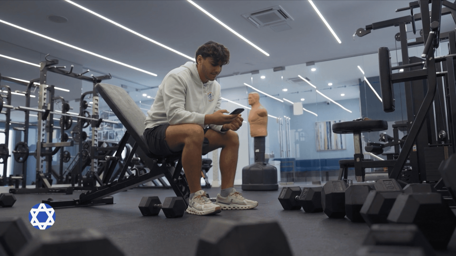Man in a white hoodie checking his phone on a weight bench in a gym