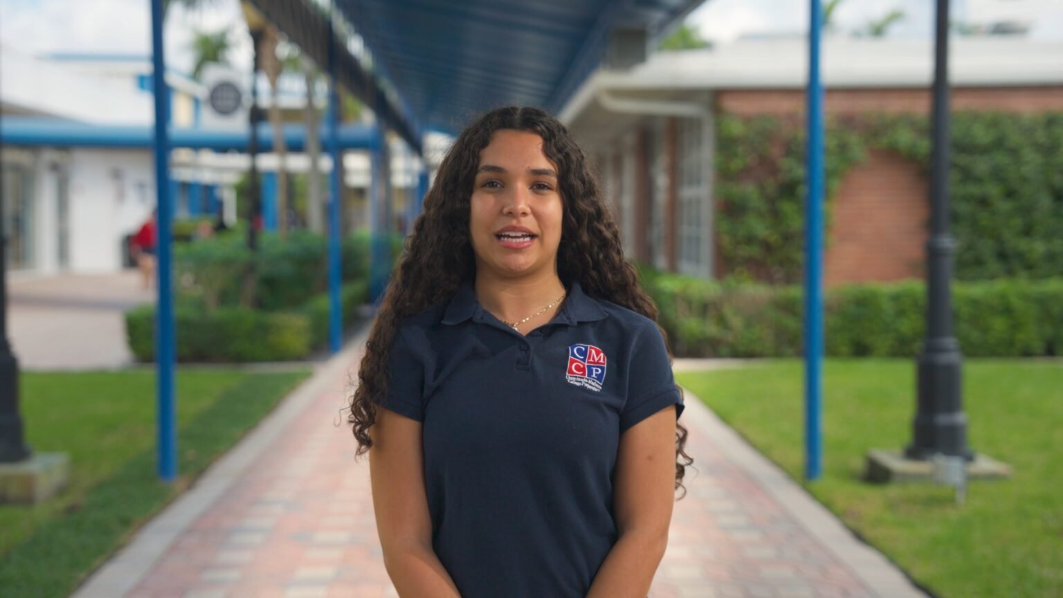 Young woman with long curly hair in a navy polo speaking on a paved walkway