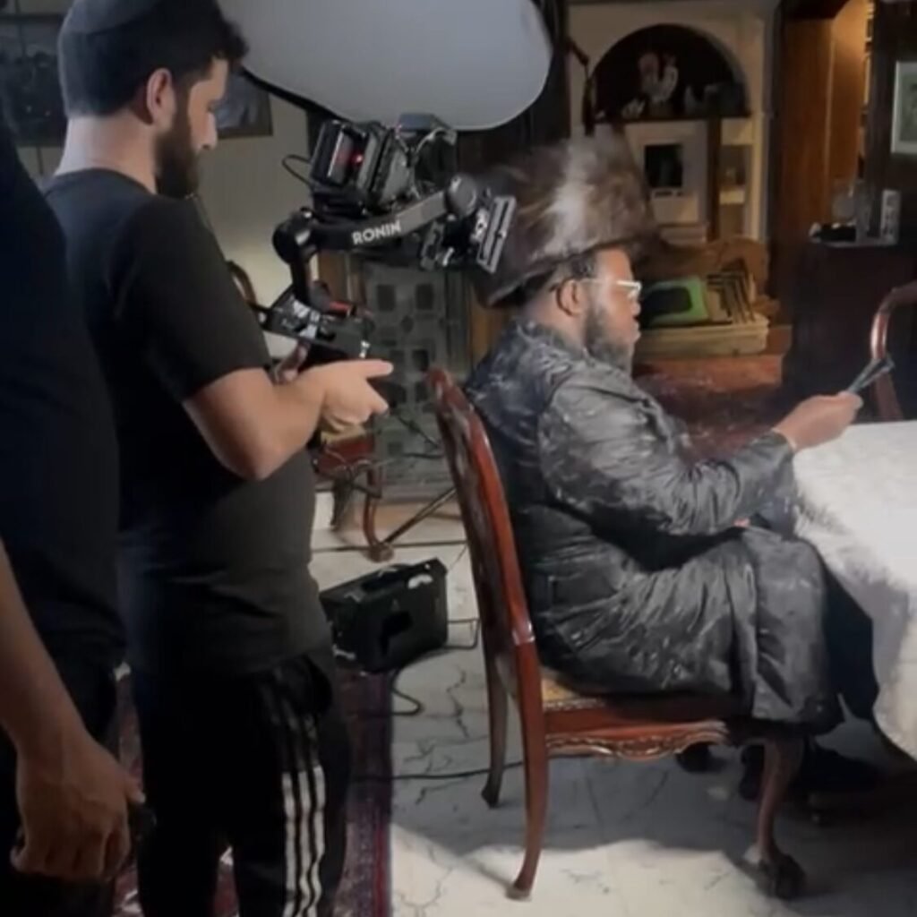 Camera operator filming a man in a fur hat and grey coat seated at a table
