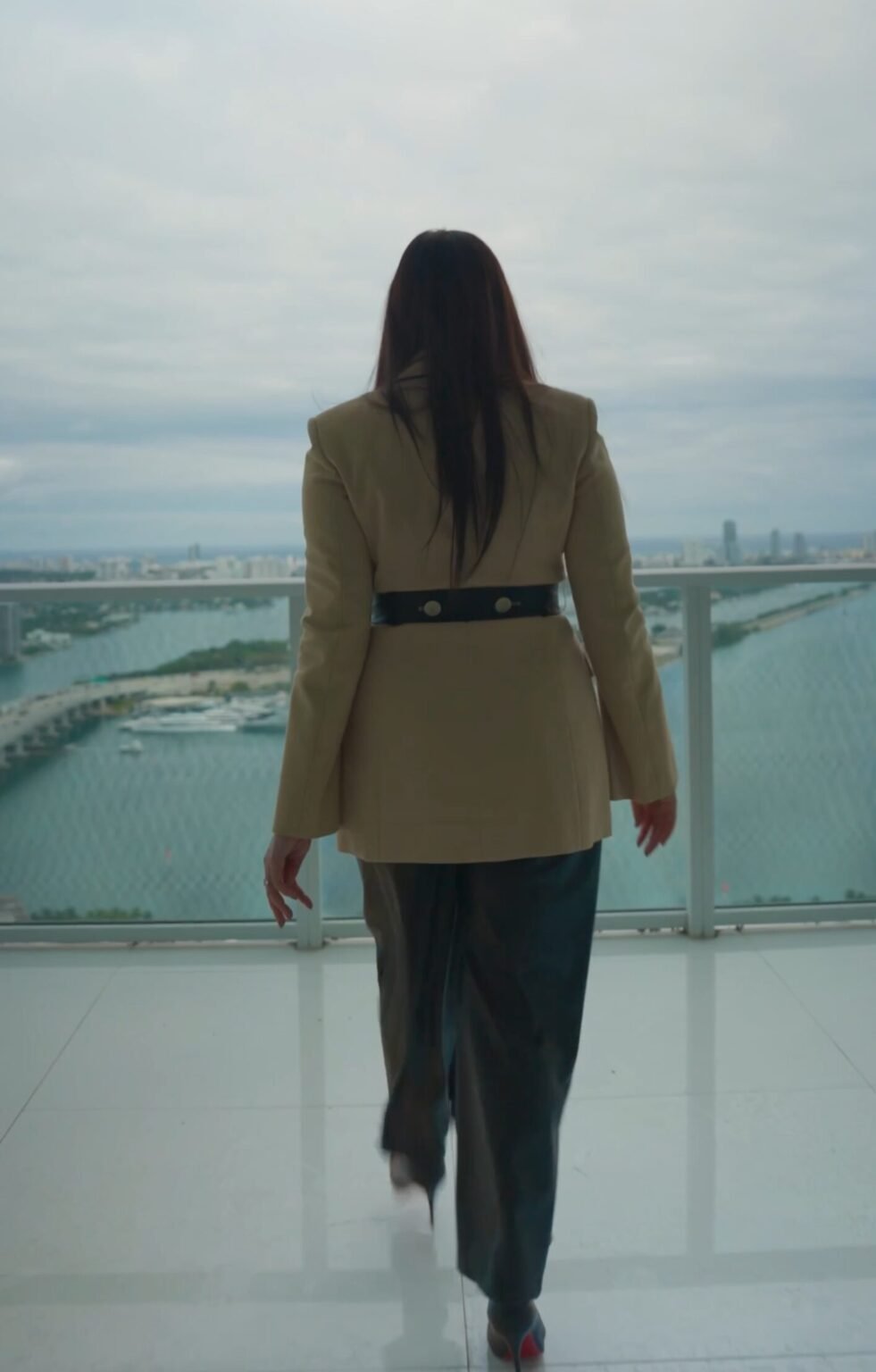Woman in a tan blazer seen from behind on a balcony overlooking water and skyline