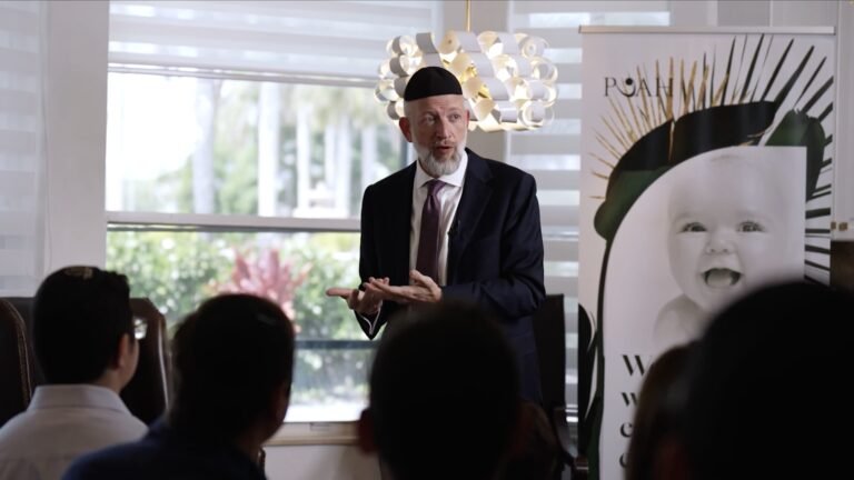 Bearded man in kippah and navy suit speaking to a seated audience beside a banner