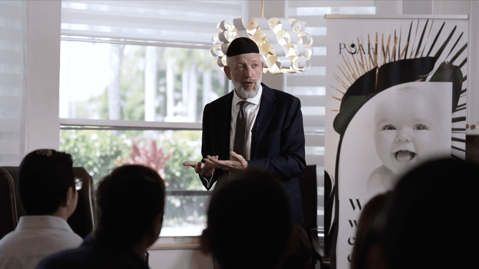 Bearded man in kippah and navy suit speaking to a seated audience beside a banner