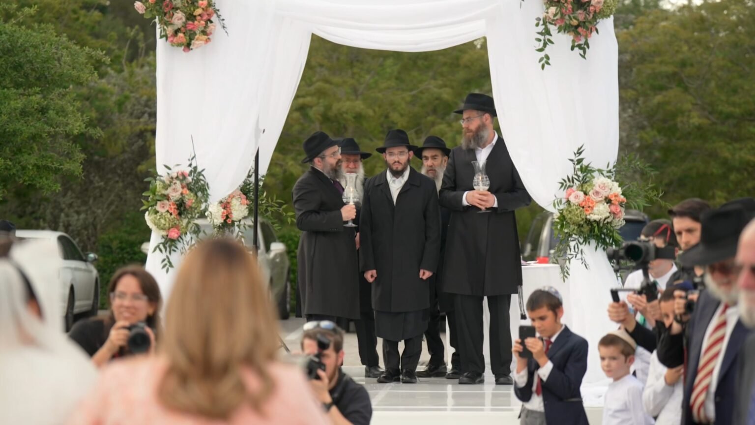 Men in black hats and coats under a white floral chuppah as guests photograph