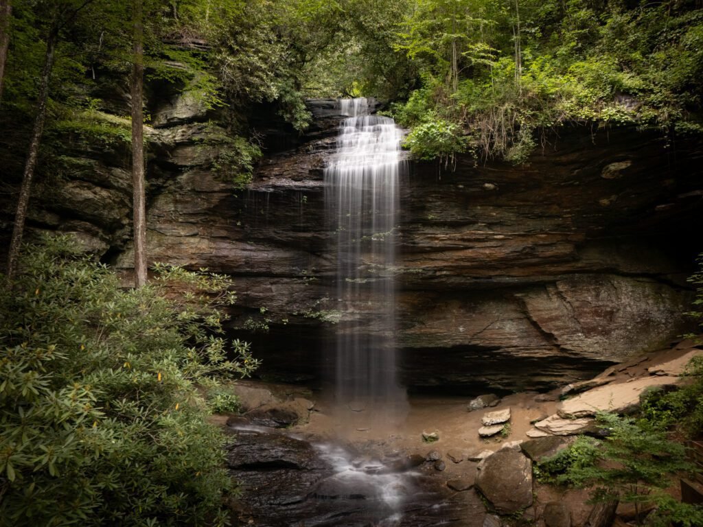 Waterfall spilling over a layered rock overhang into a shallow stream in green woods