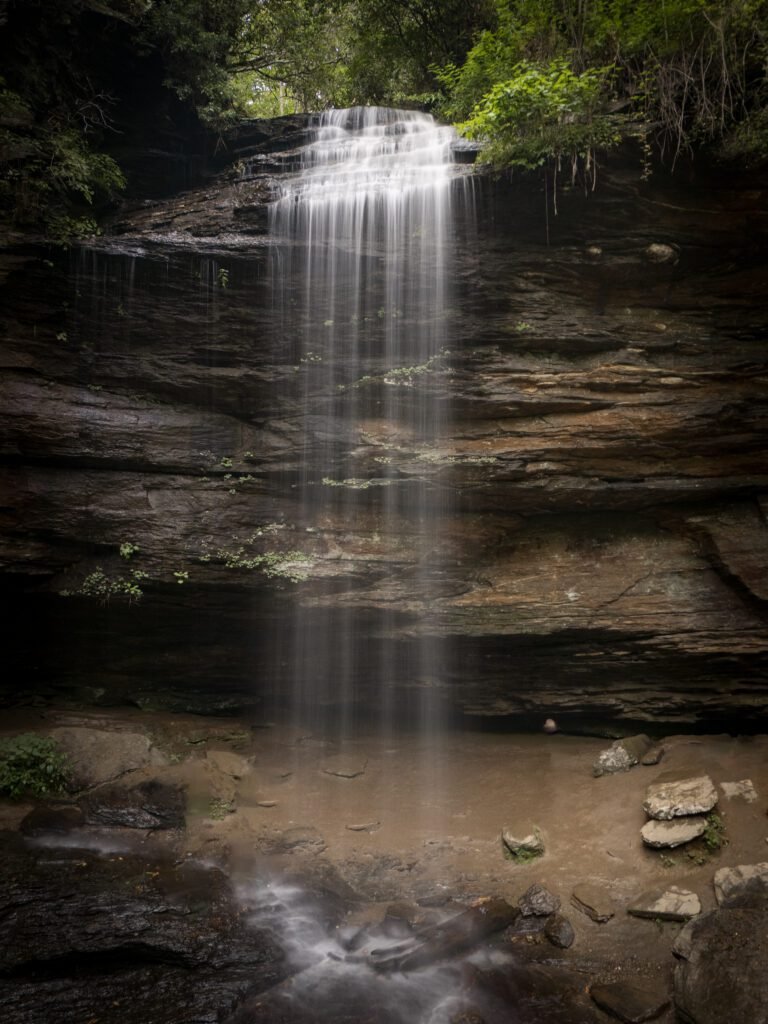 Narrow waterfall falling from a rock ledge onto sandy shallows strewn with stones