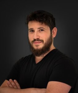 Bearded man in a black v-neck shirt with arms crossed against a dark grey backdrop