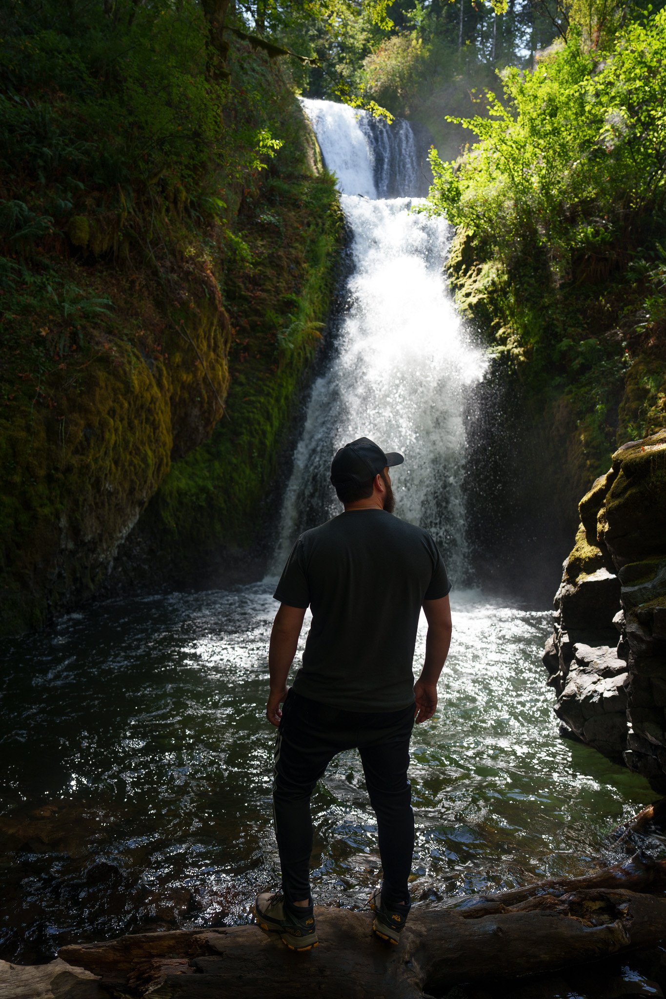 Man in a dark shirt and cap standing on a log facing a waterfall in a mossy canyon