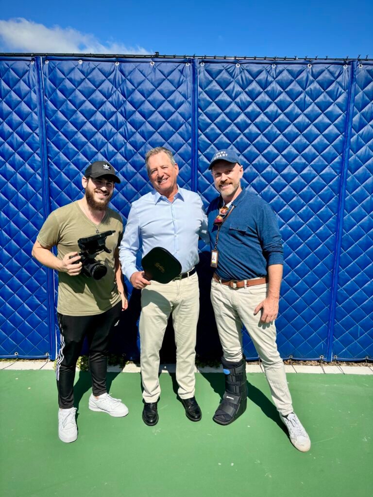 Dov Gurewicz holding a camera beside Mayor Glenn Singer and a member of the town staff on a Golden Beach court