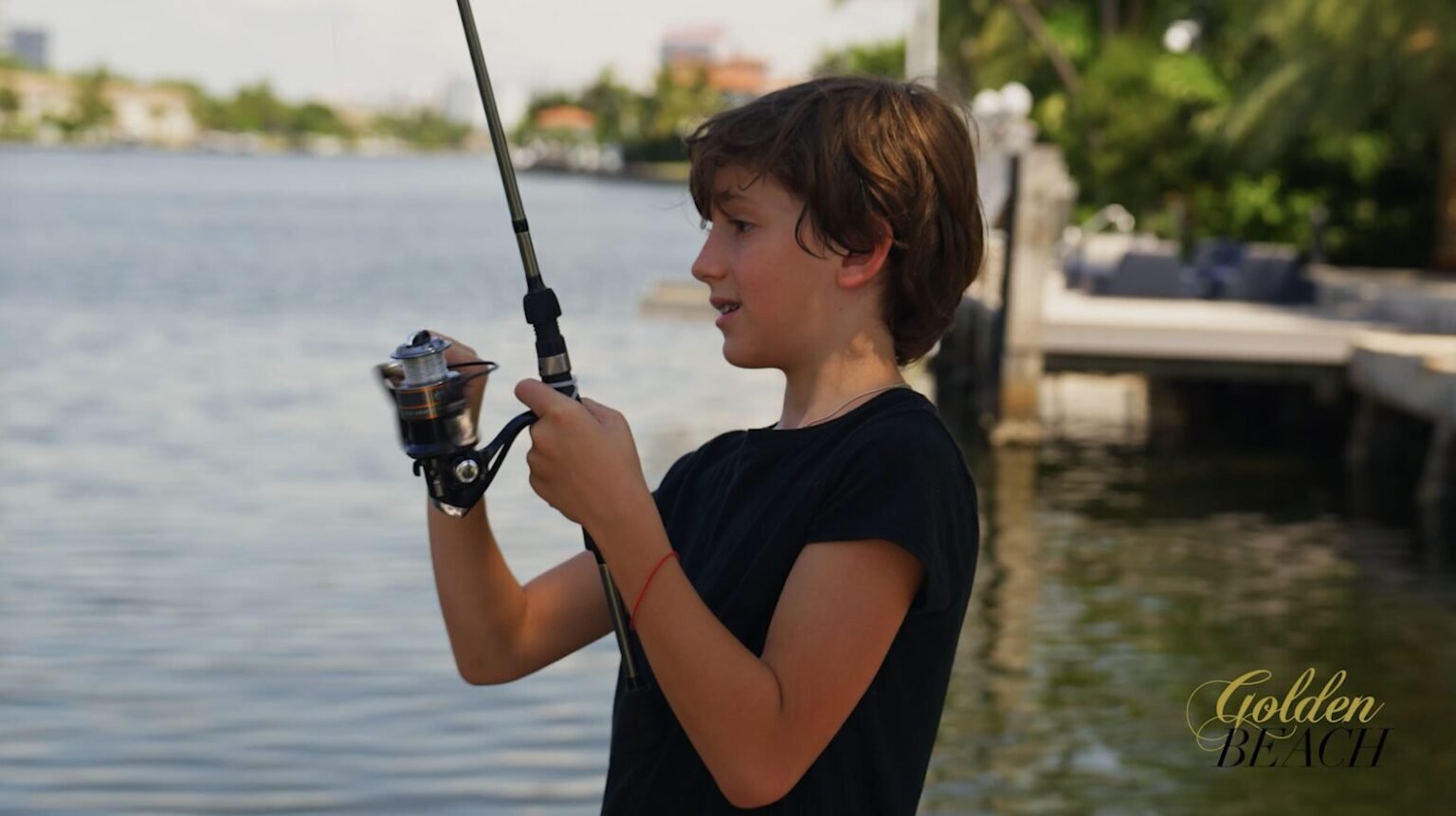 Boy in a navy t-shirt holding a fishing rod beside a waterway lined with docks