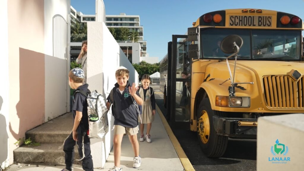 Children with backpacks walking past a yellow school bus, one boy waving