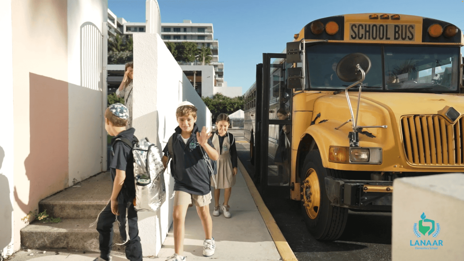 Children with backpacks walking past a yellow school bus, one boy waving