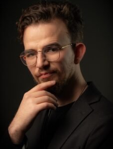 Man with curly hair and wire-rimmed glasses resting his chin on his hand, dark background