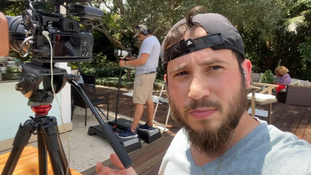 Bearded man in a backwards cap beside a tripod-mounted camera, crew filming outdoors behind him