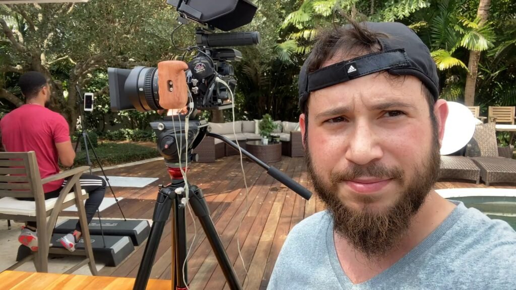 Bearded man in a backwards cap beside a tripod-mounted camera rig on a poolside deck