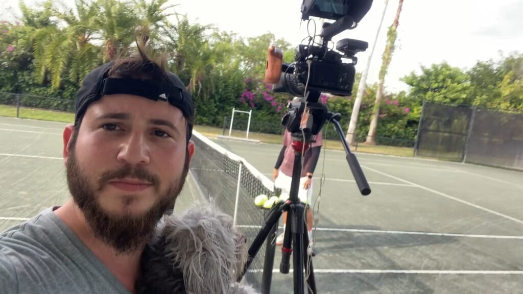 Bearded man holding a furry boom microphone on a tennis court beside a camera tripod