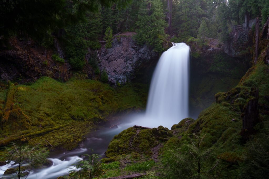 Broad waterfall dropping over a dark cliff into a river between mossy green slopes