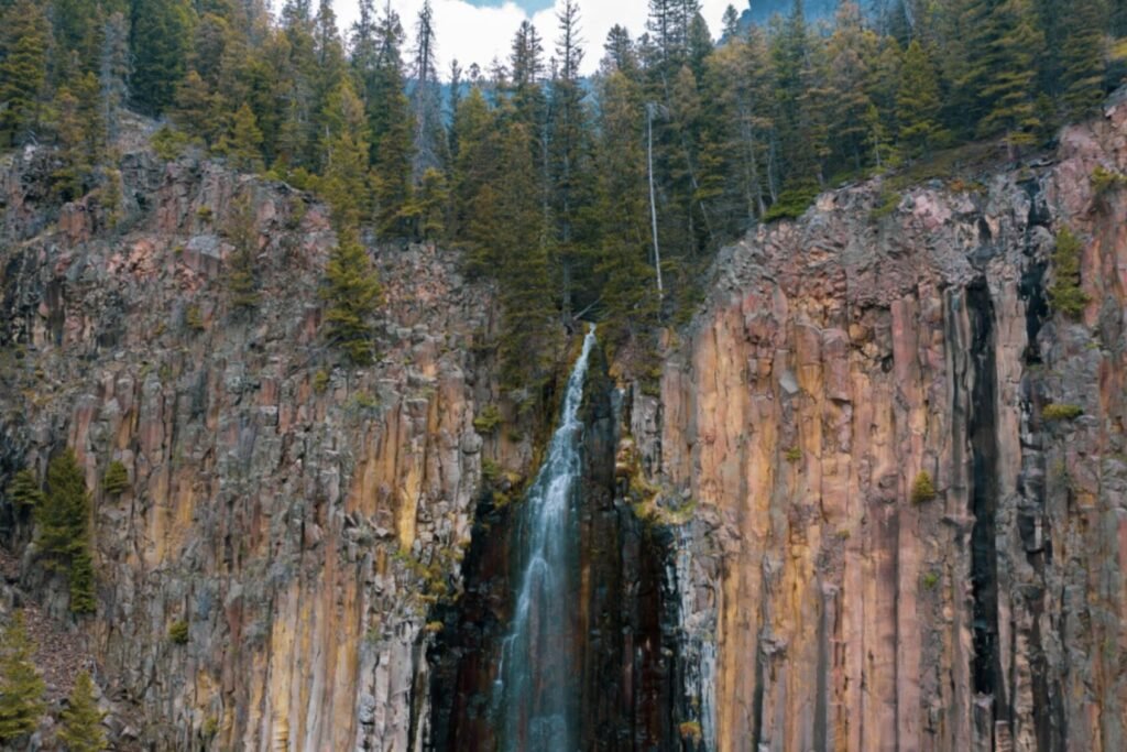 Narrow waterfall dropping between tall columnar rock cliffs topped with pine trees