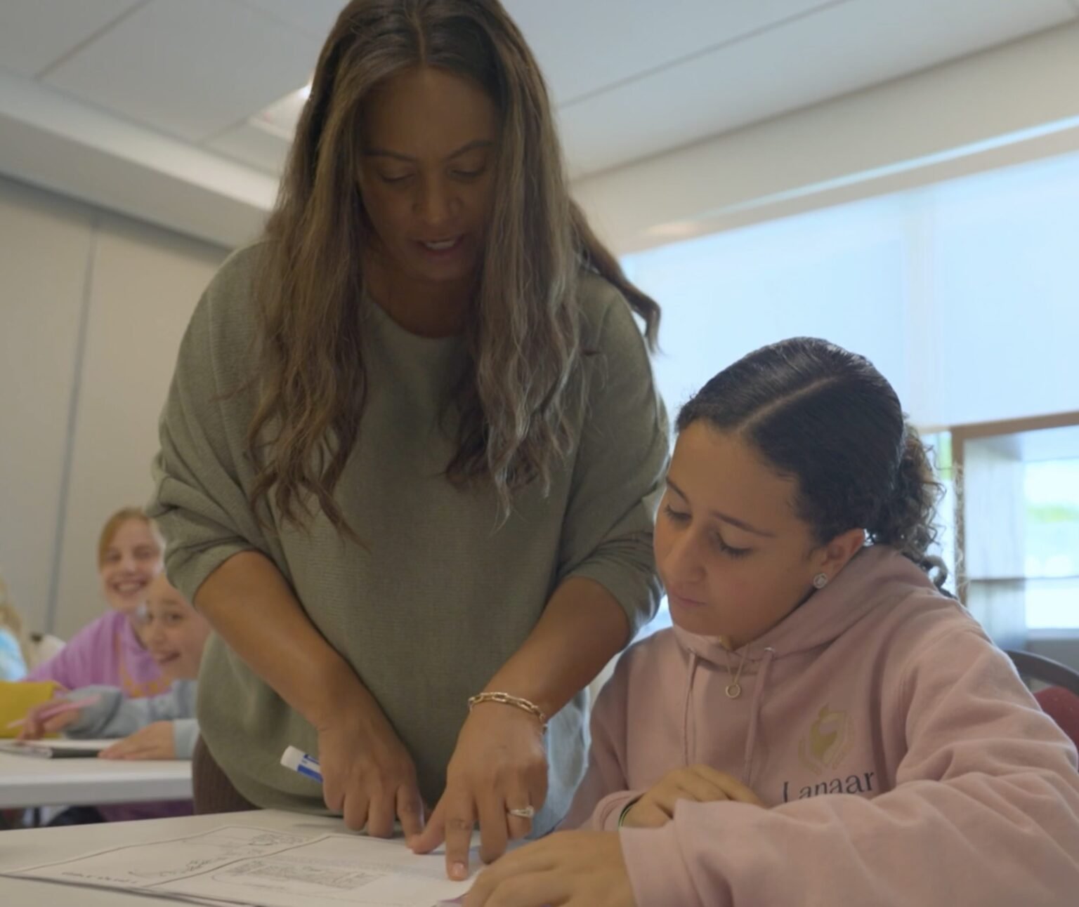 Woman leaning over a classroom desk helping a girl in a pink hoodie with worksheets