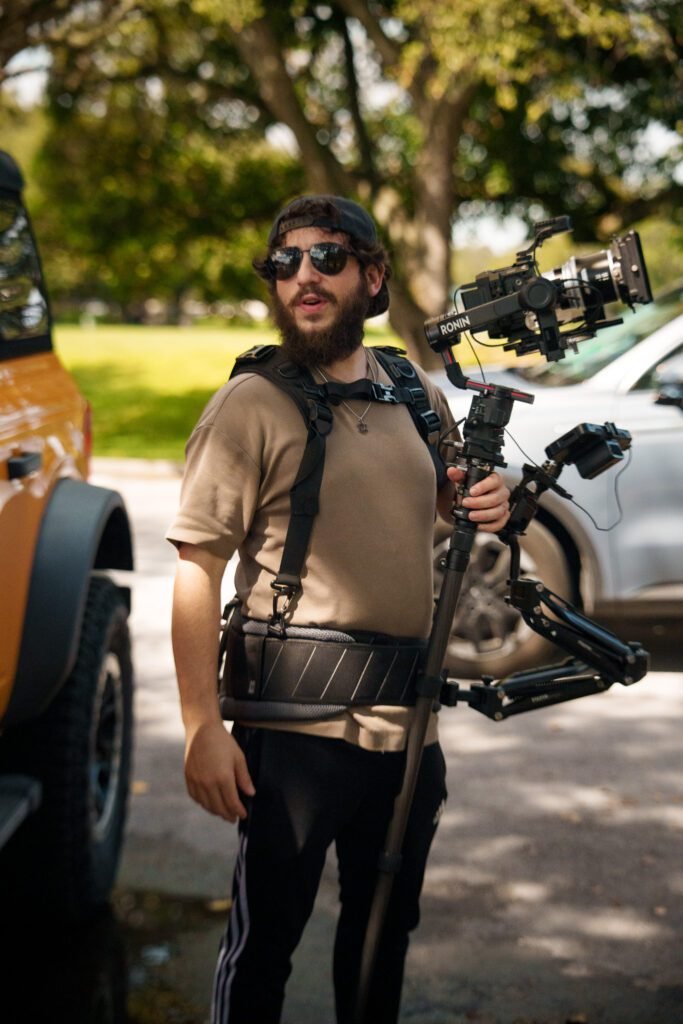 Bearded man in sunglasses wearing a camera stabilizer vest and rig beside parked cars