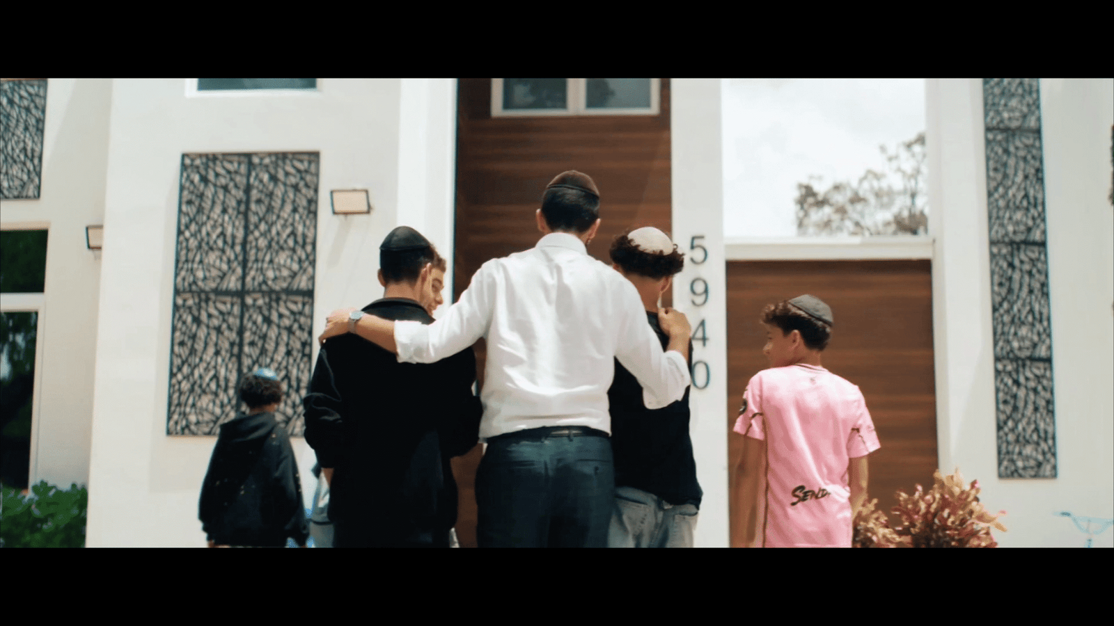 Man in a white shirt walking two boys in kippot toward a house door, girl in pink following