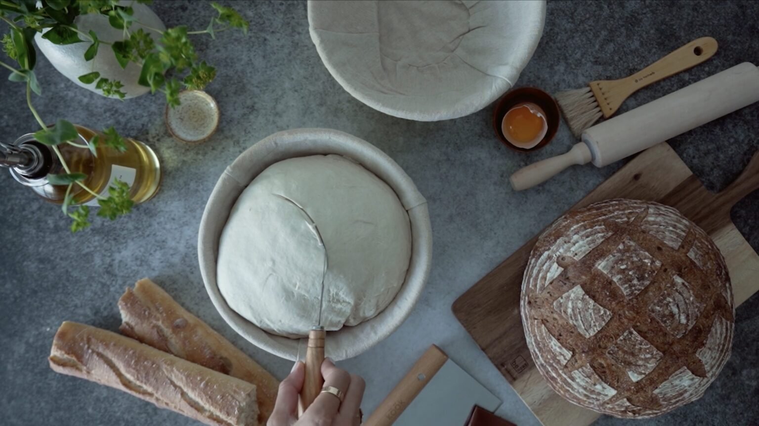 Overhead view of bread dough in proofing baskets, baguettes, scored loaf and rolling pin