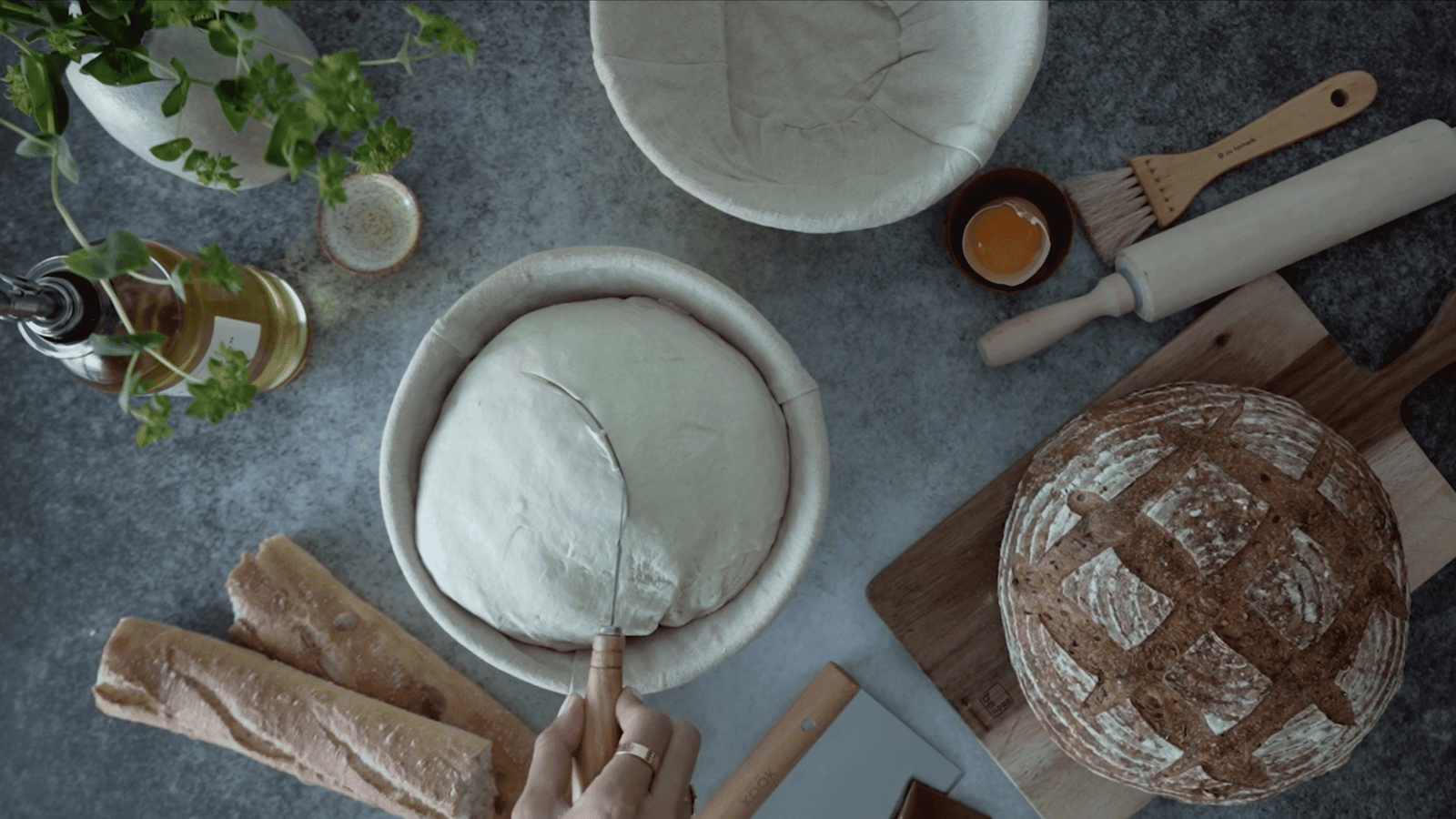 Overhead view of bread dough in proofing baskets, baguettes, scored loaf and rolling pin