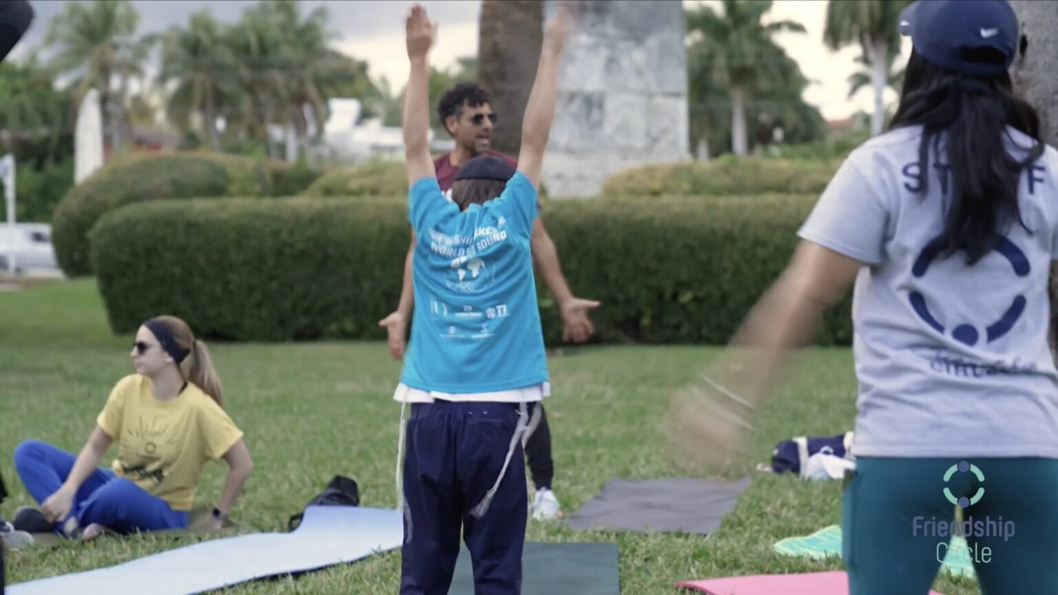 Outdoor exercise class on grass, man in a blue tank top stretching arms overhead near mats