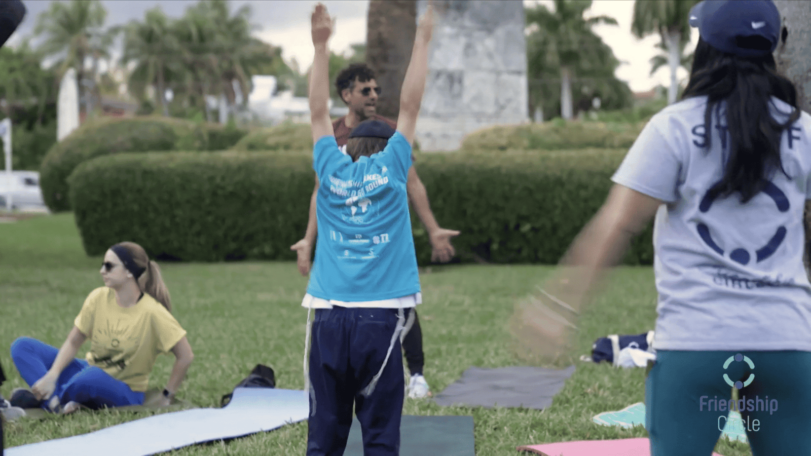 Outdoor exercise class on grass, man in a blue tank top stretching arms overhead near mats