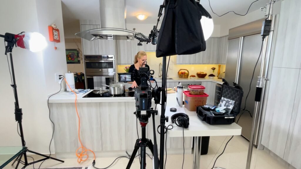 Camera on a tripod and lights aimed at a woman cooking at a kitchen island
