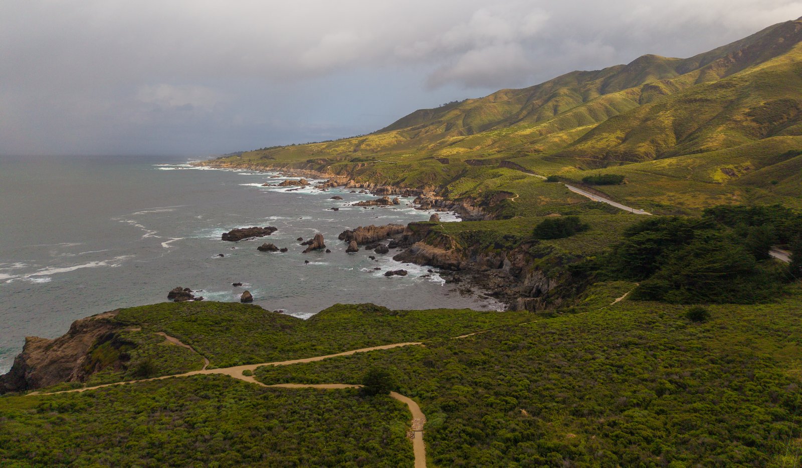 Green coastal hills above a rocky shoreline with breaking waves under overcast sky