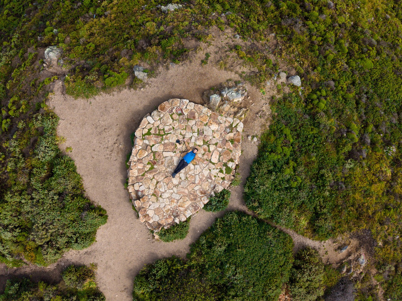 Overhead view of a person lying on a stone circle ringed by scrub and dirt paths