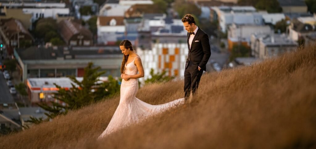 Bride and groom standing together on a grassy hillside overlooking a city at golden hour during their elopement