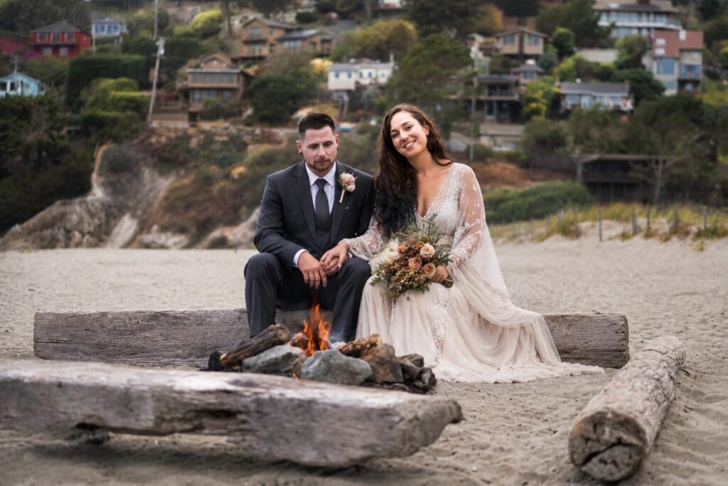 Bride and groom seated on driftwood logs beside a beach fire pit below hillside houses