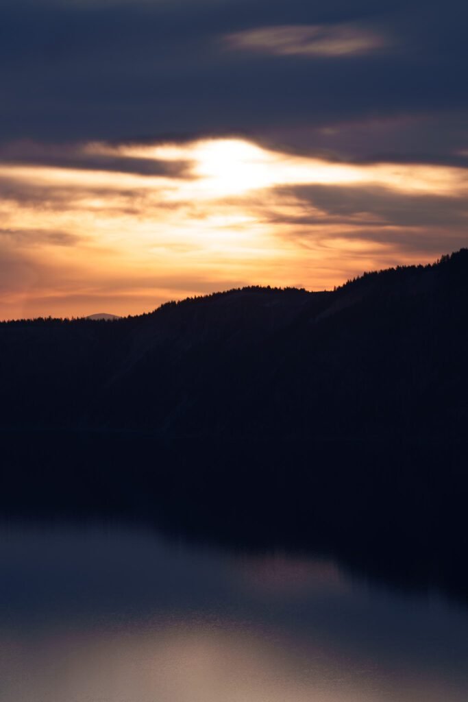 Sun breaking through clouds above a silhouetted ridge and still dark lake