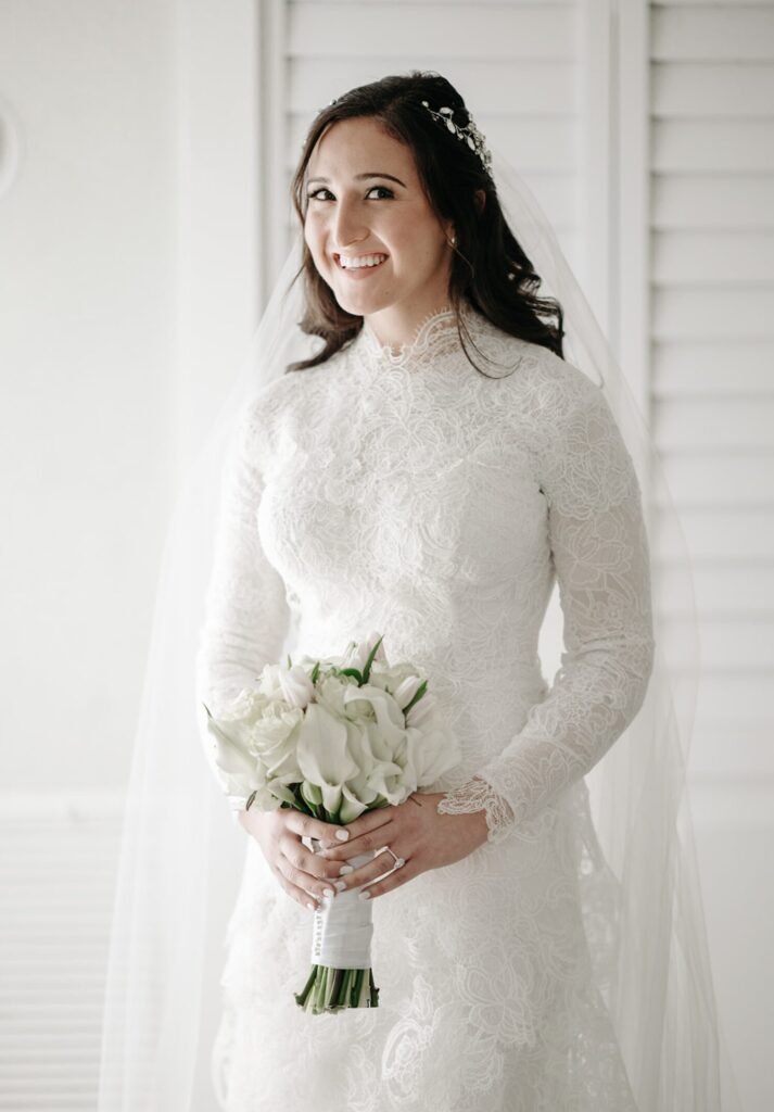 Bride in a high-neck lace gown holding calla lilies beside white louvered shutters