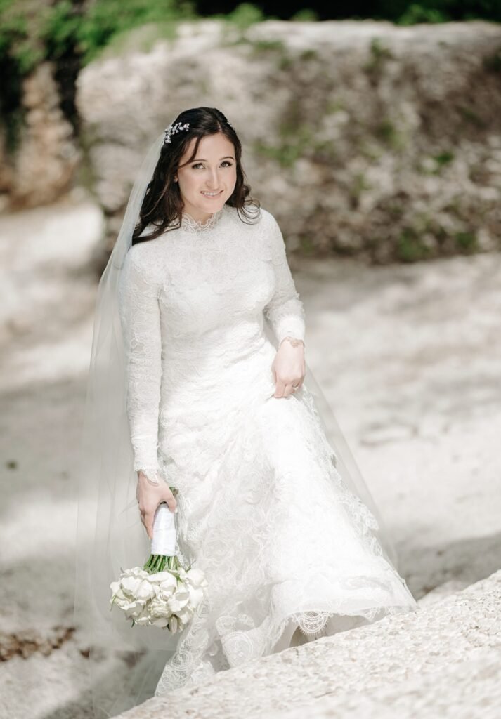 Bride in a lace gown and veil walking on pale sand holding white roses