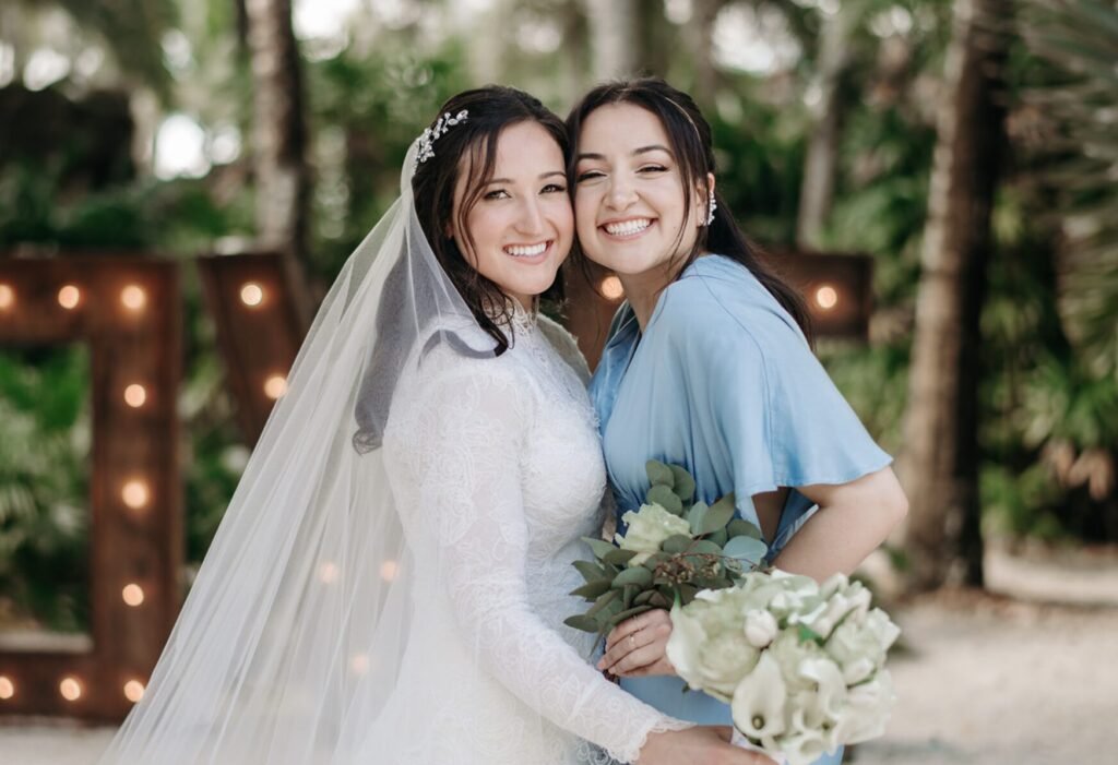 Bride and a woman in blue press cheeks together, bouquet and lit marquee letters behind