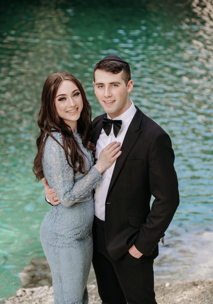 Woman in a pale blue lace gown beside a man in a tuxedo by turquoise water