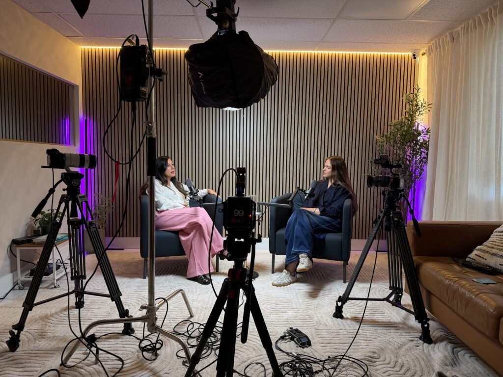 Interview set with two women in armchairs framed by three tripod cameras and overhead light