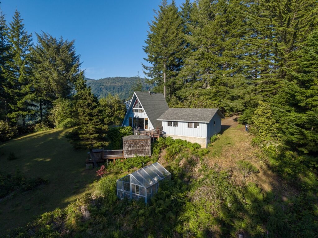 White gabled house on a grassy hillside among fir trees, greenhouse below deck