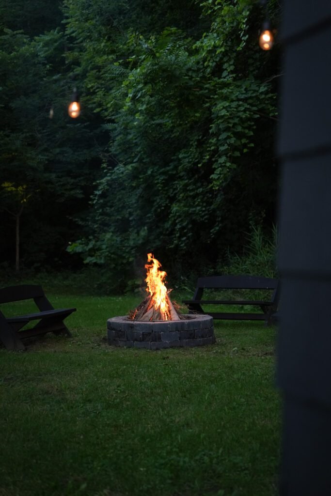 Campfire burning in a stone fire pit at dusk with string lights above and benches around it