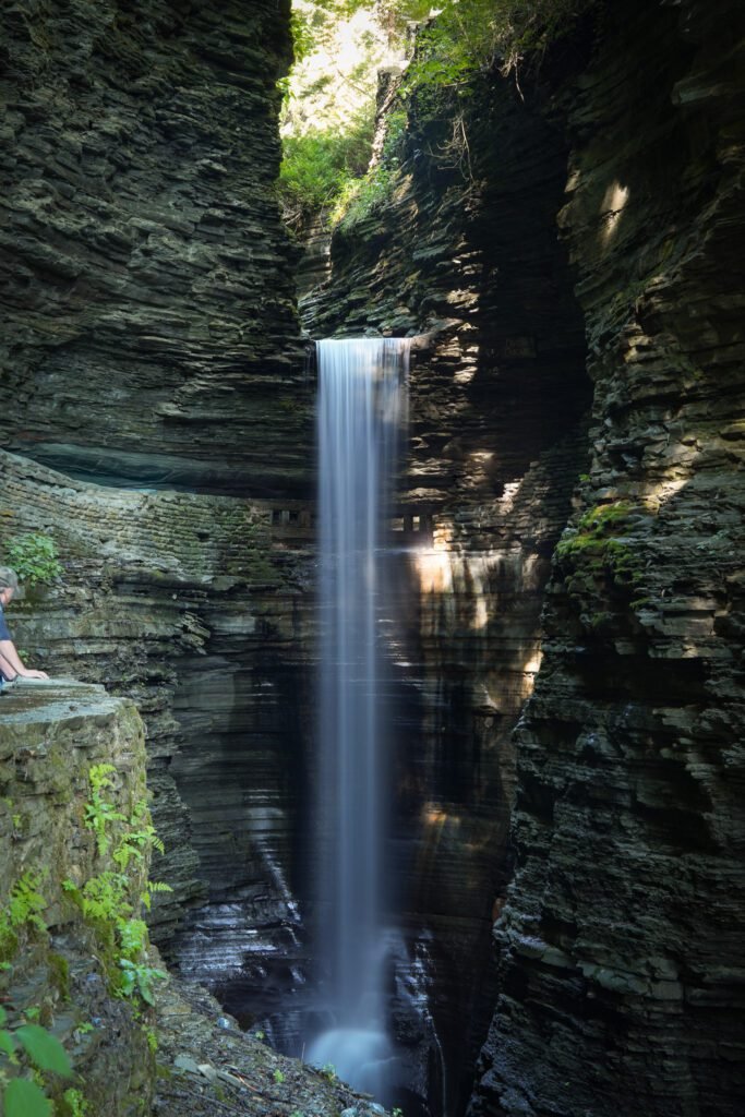 Rainbow Falls at Watkins Glen State Park, a tall veil of water falling into the shale gorge with a hiker on the ledge beside it